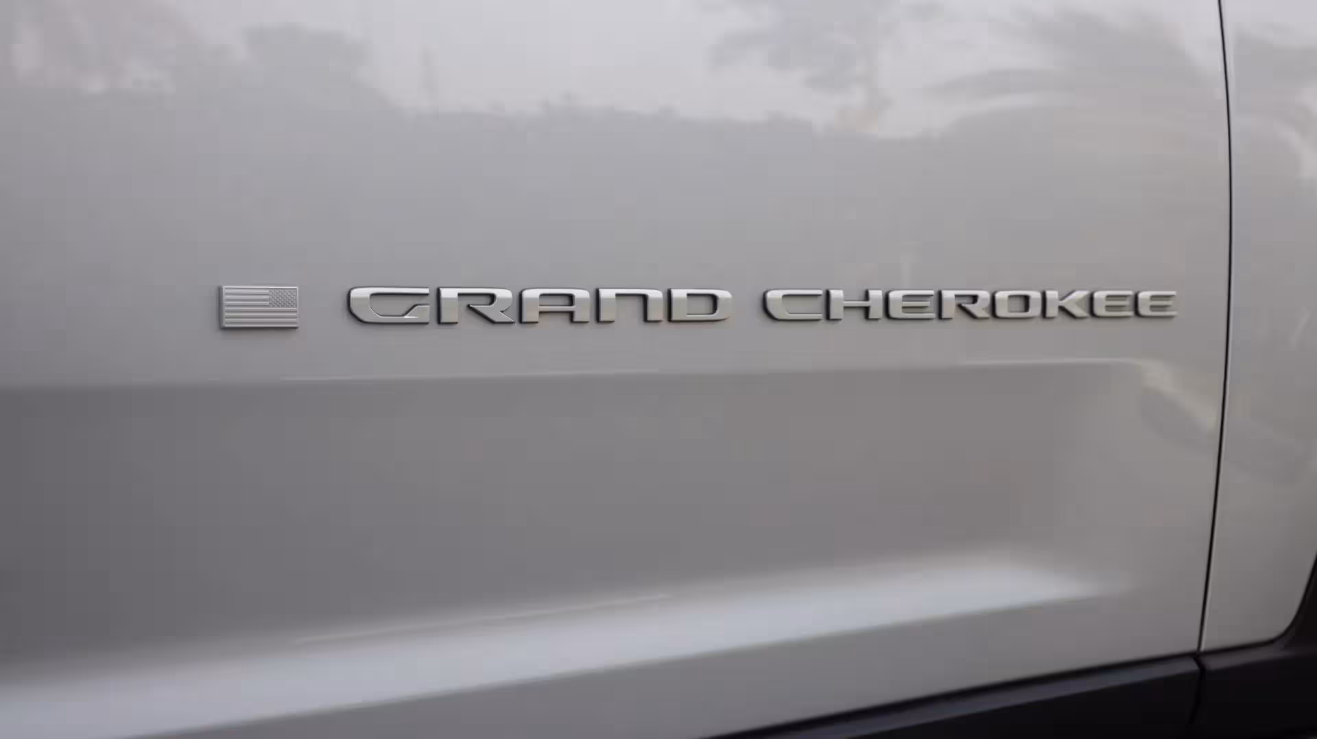Close-up of a perforated metallic side step and 'GRAND CHEROKEE' badge on the lower side of a silver Jeep Grand Cherokee.