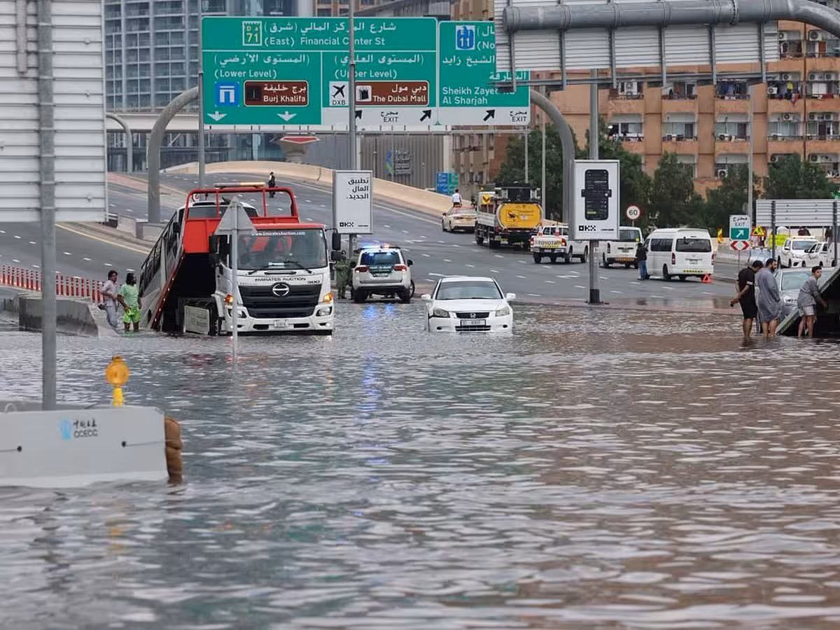 A dark grey SUV and several other vehicles driving through deep floodwater on a city street, creating large splashes. Buildings are visible in the background under an overcast sky.