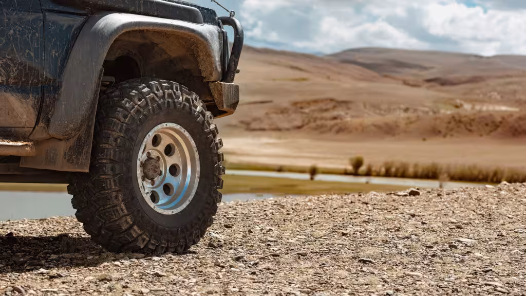 A dark SUV equipped with robust all-terrain tires and silver alloy wheels, parked on a gravelly desert terrain with an arid, hilly landscape in the background, signifying off-road adventure.