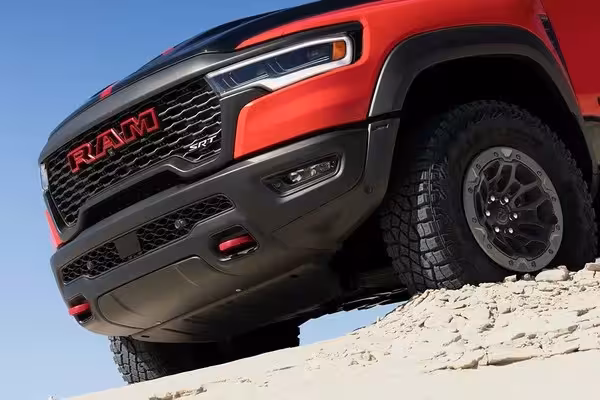 Low-angle front view of a red 2027 Ram 1500 SRT TRX perched on a sandy dune, highlighting its aggressive front fascia, large off-road tires, and prominent red tow hooks.