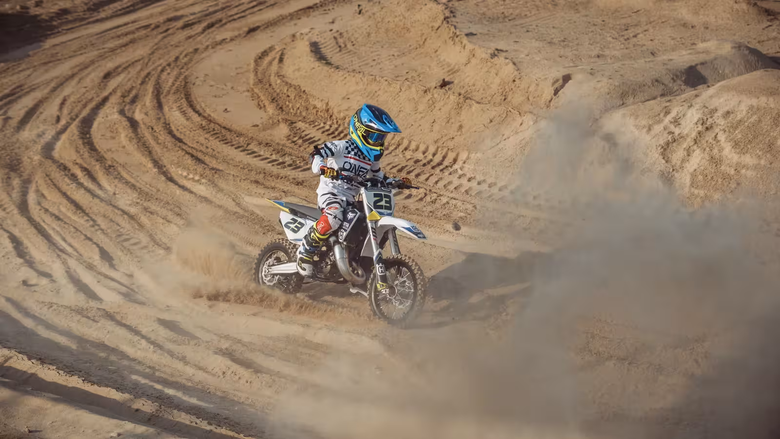 Motocross rider in black and neon yellow gear on a white and blue dirt bike, leaning into a turn and kicking up sand on the DMX track.