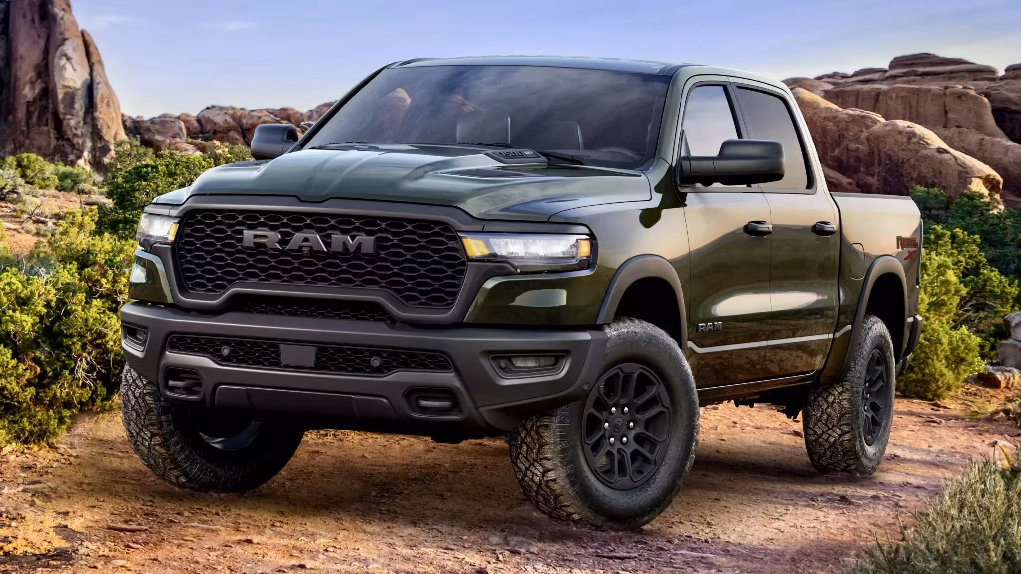 The 2026 Ram 1500 Rebel X Crew Cab 4x4 in the new Tank Clear-Coat color, showcasing its aggressive off-road stance and black wheels against a desert landscape.
