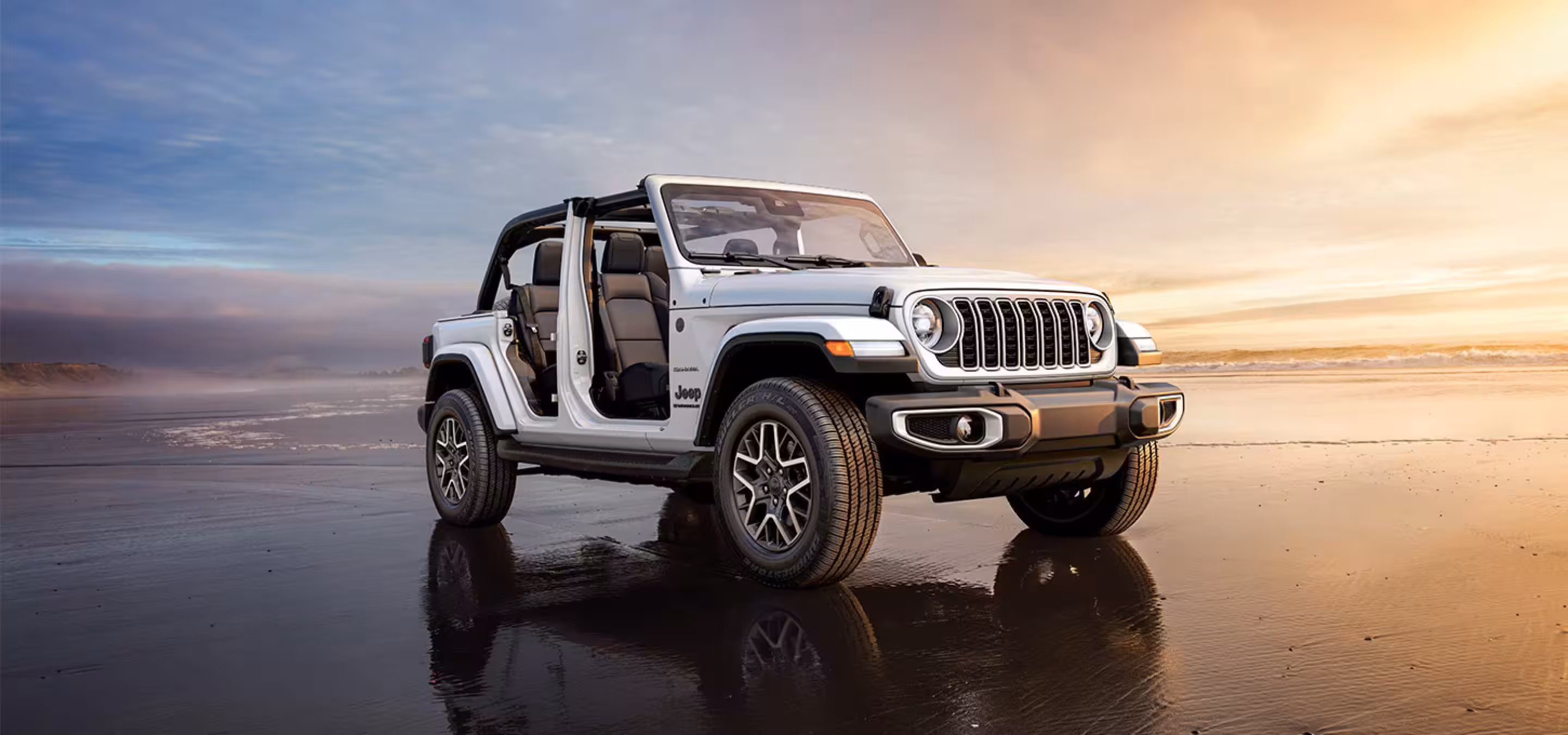 White 2026 Jeep Wrangler two-door model with doors removed and soft top down, parked on a wet beach at sunset.