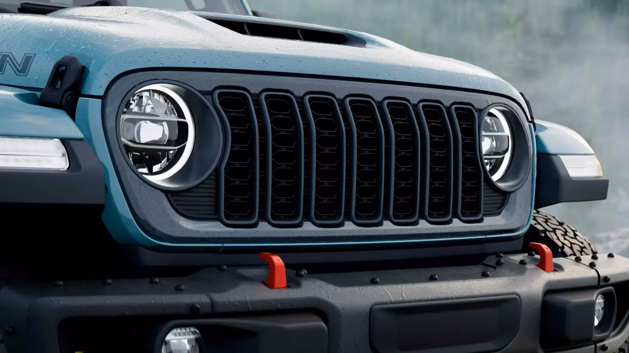 Close-up of the 2026 Jeep Wrangler Rockslide's front grille and LED headlights in Earl color, showing the dark Anvil grille and red tow hooks.