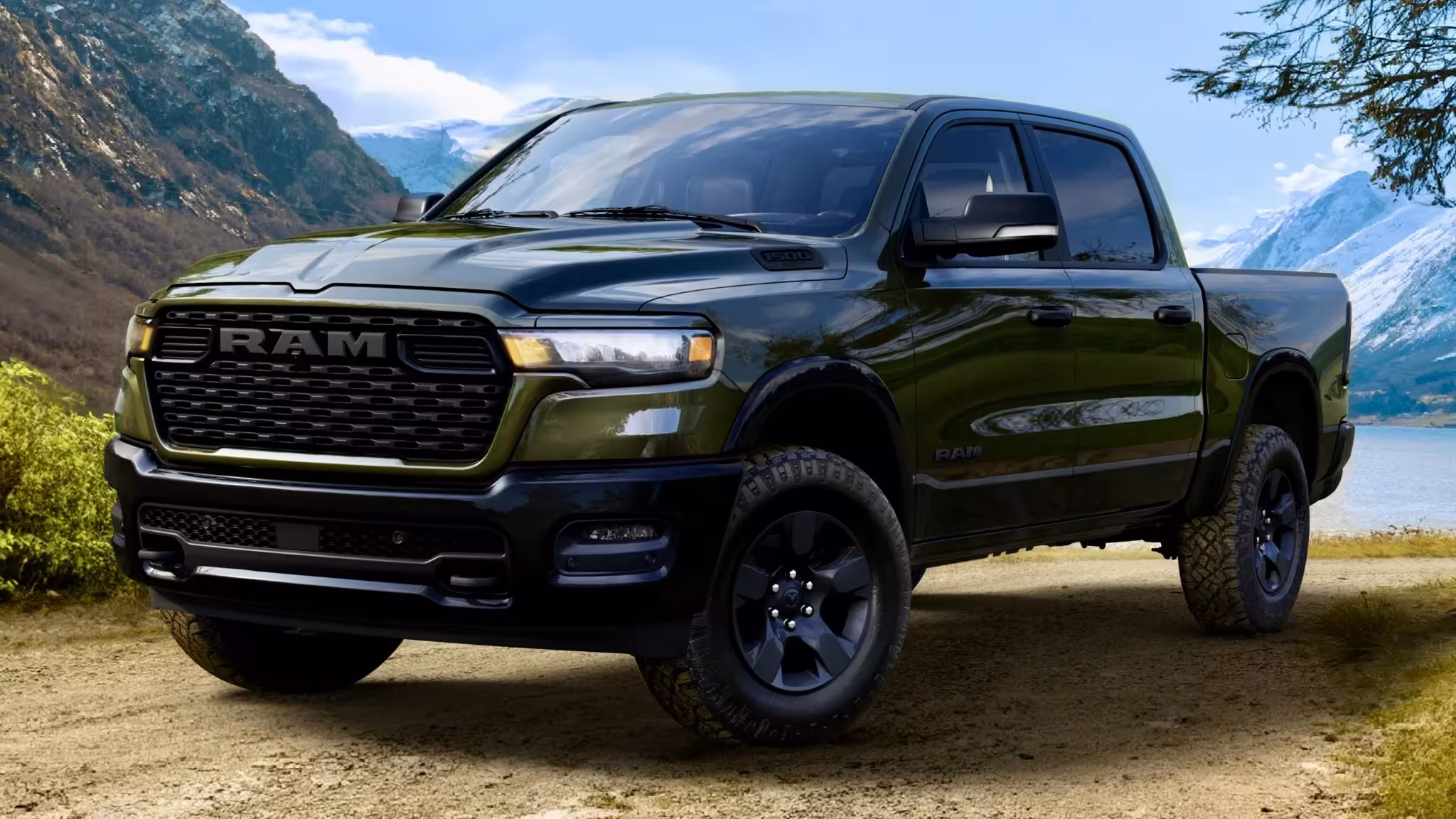 Olive green 2026 Ram 1500 BackCountry on a dirt path by a lake with snowy mountains in the background, showcasing its front end and stance.