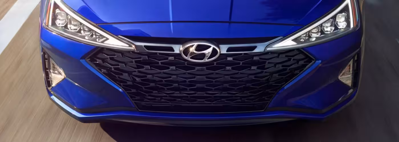 The front grille of a vibrant blue Hyundai car, featuring the prominent silver 'H' logo in the center of a hexagonal mesh pattern, flanked by modern headlights.