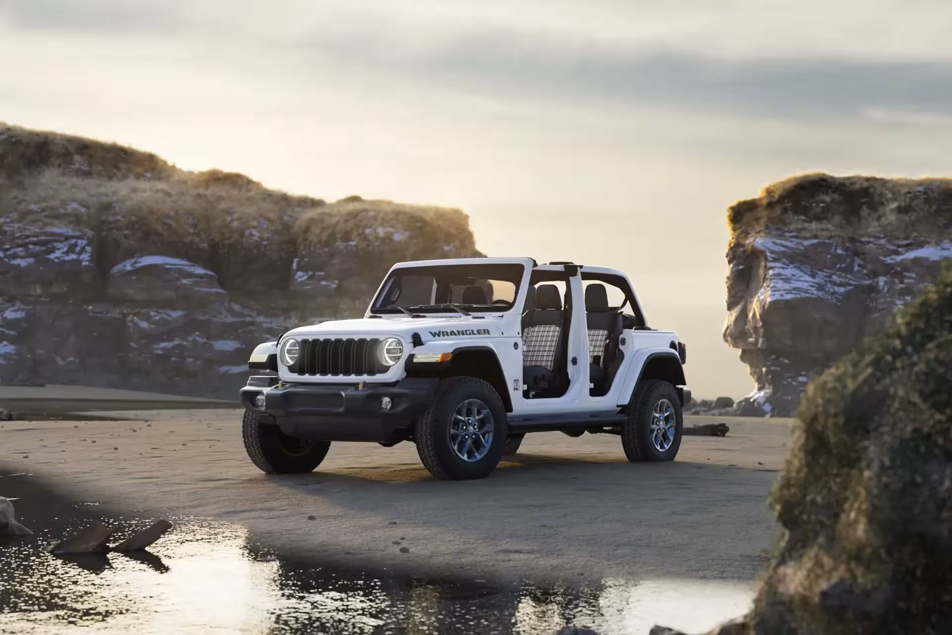 A white Jeep Wrangler 85th Anniversary Special Edition, with doors and roof panels removed, parked on a sandy beach next to a small pool of water, with rugged cliffs in the background at sunrise.