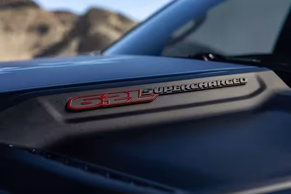 Wide shot of a dark grey 2027 Ram 1500 SRT TRX driving aggressively on a dirt road, kicking up a plume of dust, showcasing its powerful off-road presence.
