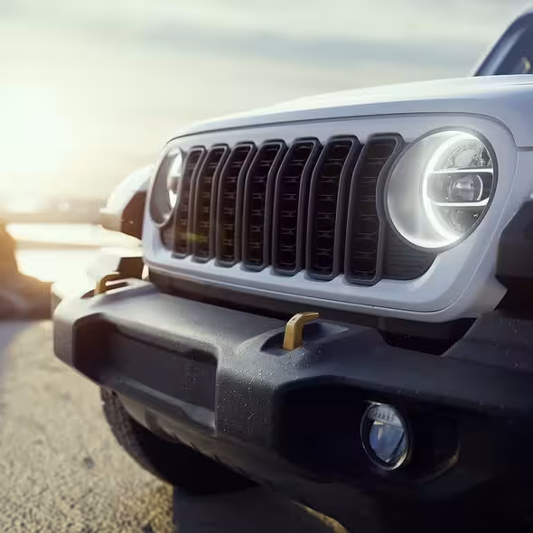 Close-up of a white Jeep Wrangler's front grille and round LED headlights, featuring gold-colored tow hooks on the bumper, against a bright, sunny background.