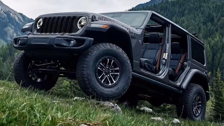 2027 Jeep Wrangler Rocky Mountain 392 off-roading on a rocky, grassy incline with mountains in the background, showcasing its 35-inch BFGoodrich tires and open doors.