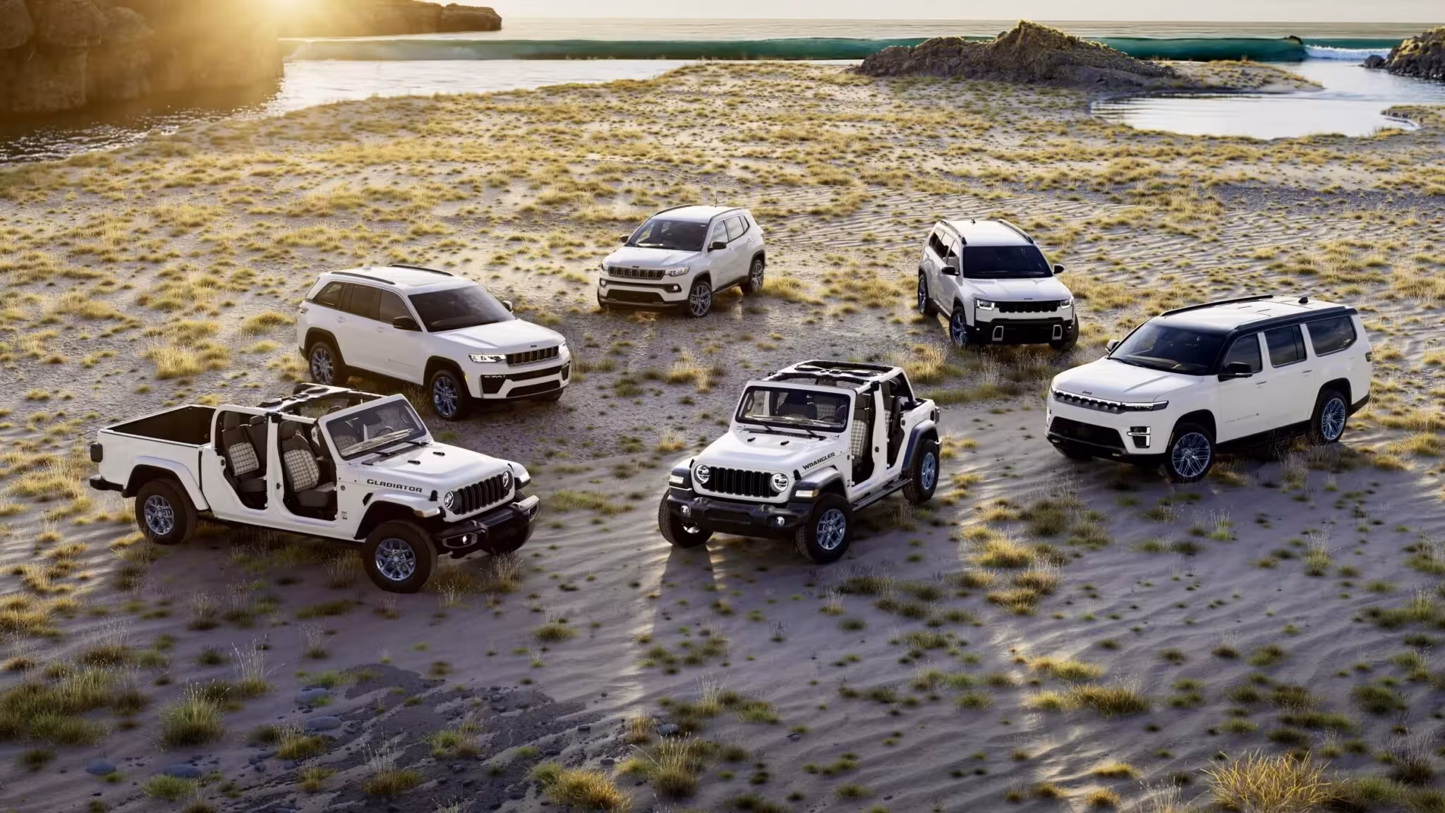 Six white Jeep 85th Anniversary Special Edition vehicles parked on a sandy beach at sunrise, including a Wrangler, Gladiator, Grand Cherokee, Grand Wagoneer, Compass, and potentially a Cherokee.
