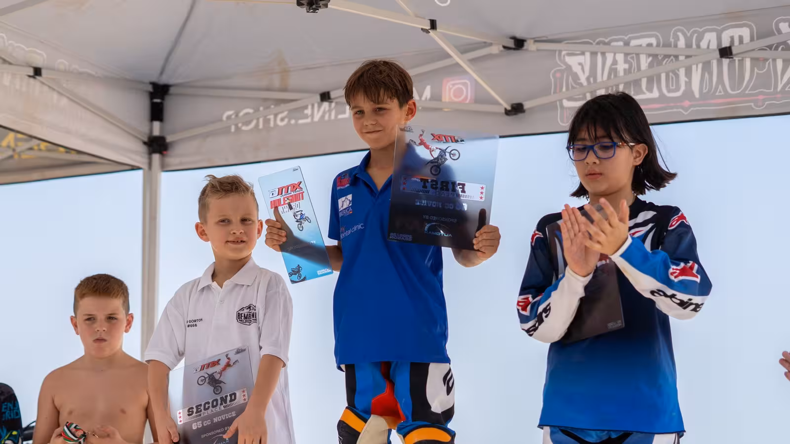 Three young participants on a podium, with the center boy holding a 'FIRST' place award, celebrating their achievements at the DMX Junior Championship.