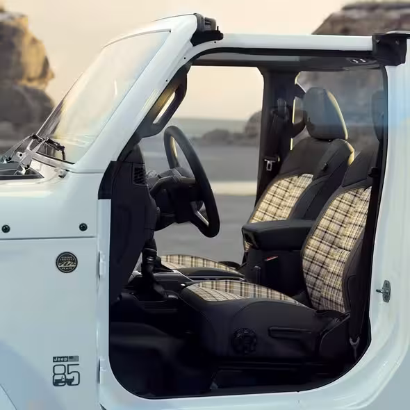 Open door view of a white Jeep Wrangler 85th Anniversary Special Edition, showing the plaid interior seats, 'Jeep 85' door logo, and fender badge, set against a bright outdoor background.