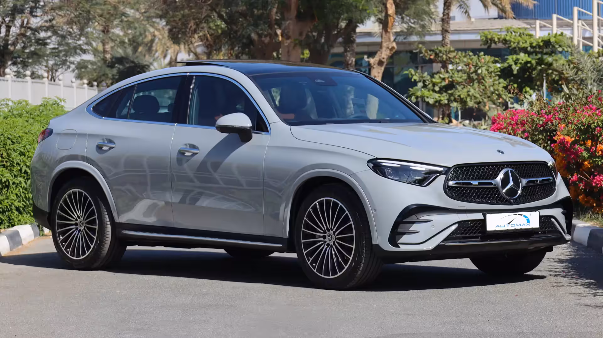 Side profile and front three-quarter view of a light grey 2026 Mercedes-Benz GLC 200 Coupe, showcasing its sleek sloping roofline, black panoramic sunroof, and stylish multi-spoke alloy wheels.
