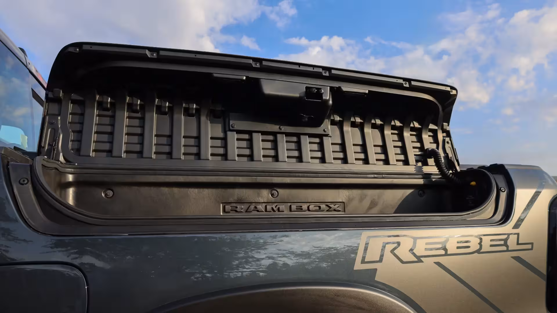 Front three-quarter view of an olive green 2026 Ram 1500 Rebel on a paved road in a desert environment, showcasing its off-road tires and black wheels.