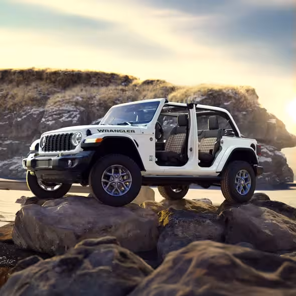 A white Jeep Wrangler 85th Anniversary Special Edition with doors and roof removed, parked dynamically on large rocks on a beach with rugged cliffs in the background at sunset.