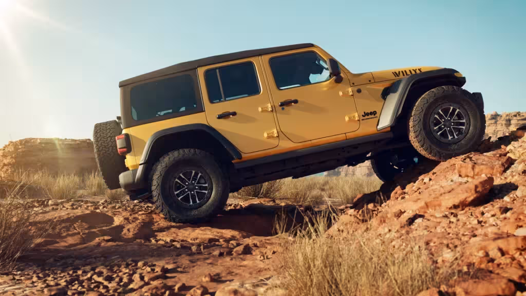 Side profile of the 2026 Jeep Wrangler Unlimited Willys 392 in Goldilocks, expertly rock crawling over a challenging terrain. The 'WILLYS' decal on the hood and large BFGoodrich KO2 tires are prominent.