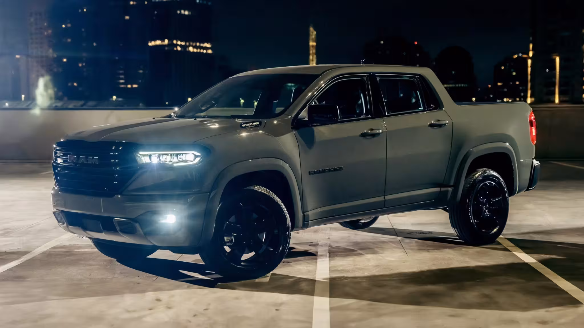 Side profile view of the 2027 Ram Rampage Laramie Night Edition in a dark gray metallic finish, with illuminated headlights and fog lights, parked at night with city lights in the background.