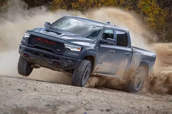 Close-up of the front grille of a black 2027 Ram TRX, featuring red 'RAM' lettering, a functional hood scoop with amber LED identification lamps, and a bold red racing stripe.