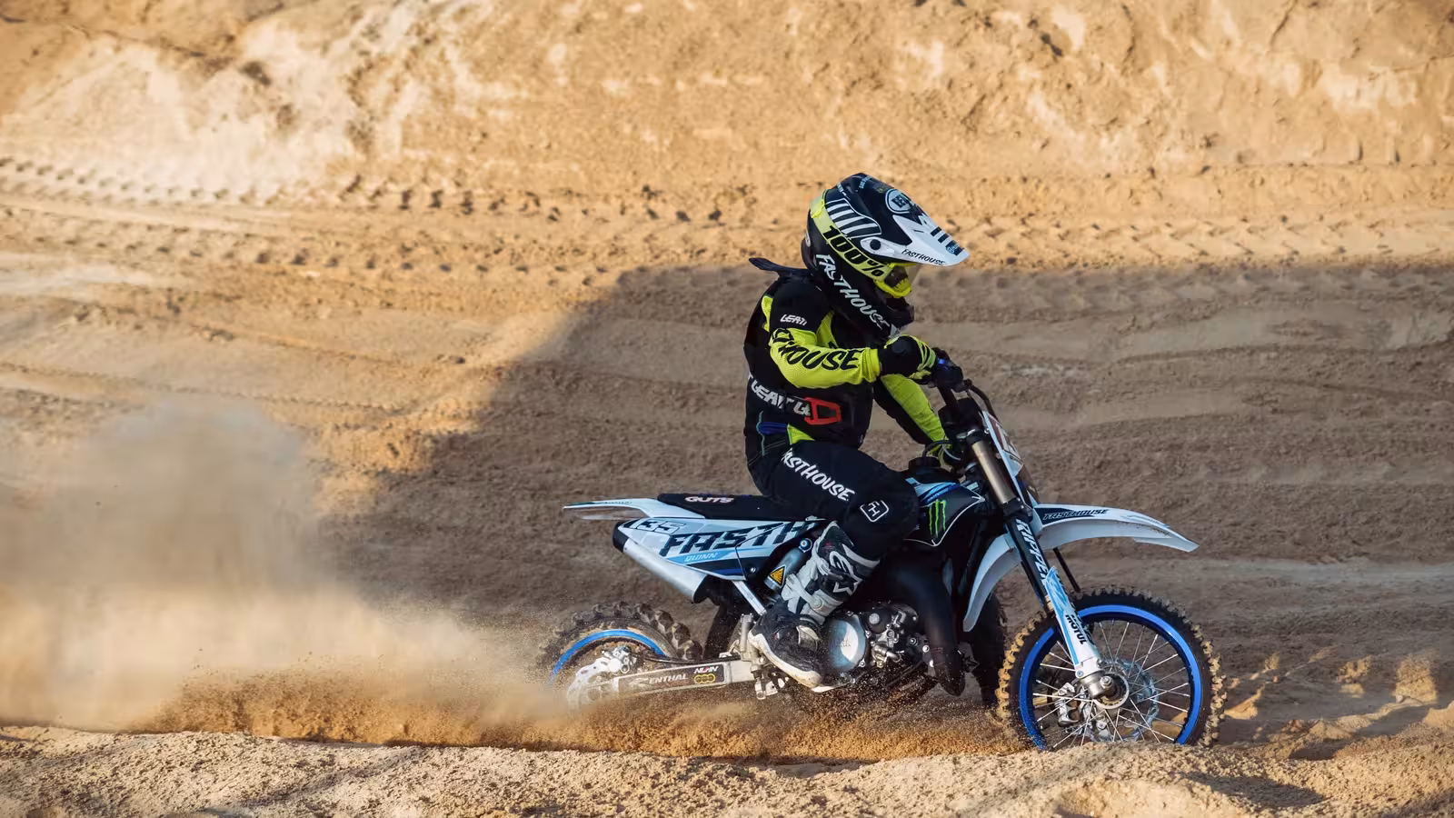 Young motocross rider, number 27, in vibrant gear, making a sharp turn on a sandy DMX track, kicking up a large spray of sand.