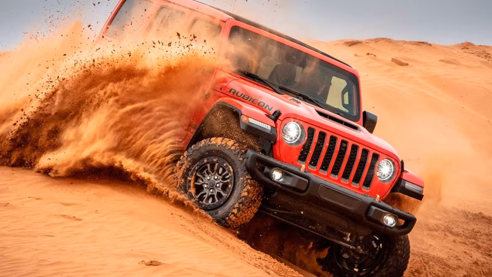 A vibrant red Jeep Wrangler Rubicon aggressively driving up a steep sand dune, with sand spraying from its wheels, demonstrating extreme off-road capability in the desert.