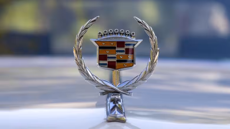 Close-up of a classic Cadillac shield logo, an ornate metallic emblem featuring a detailed crest with multiple colored sections, a small crown at the top, and a surrounding laurel wreath.